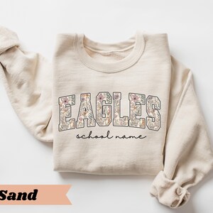 May include: A sand-colored sweatshirt with the word "EAGLES" in a floral design across the chest. Below the word "EAGLES" is the text "school name" in a cursive font. The sleeves are rolled up.