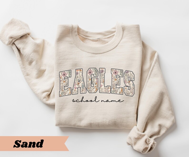 May include: A sand-colored sweatshirt with the word "EAGLES" in a floral design across the chest. Below the word "EAGLES" is the text "school name" in a cursive font. The sleeves are rolled up.