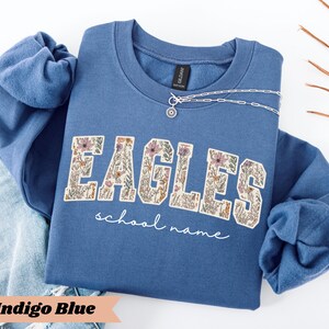 May include: Indigo blue sweatshirt with the word "EAGLES" in a floral design. The words "school name" are written in a script font below the word "EAGLES". A silver necklace with a circular pendant is on the sweatshirt.
