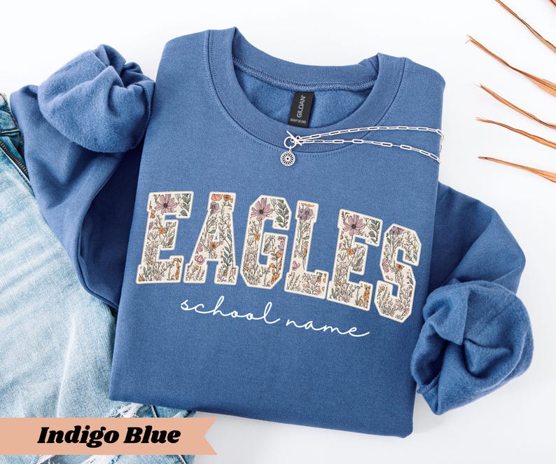 May include: Indigo blue sweatshirt with the word "EAGLES" in a floral design. The words "school name" are written in a script font below the word "EAGLES". A silver necklace with a circular pendant is on the sweatshirt.
