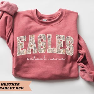 May include: Heather scarlet red sweatshirt with the word "EAGLES" in a floral design. Below the word "EAGLES" is the phrase "school name" in a cursive font. The sweatshirt has long sleeves and a crew neck.