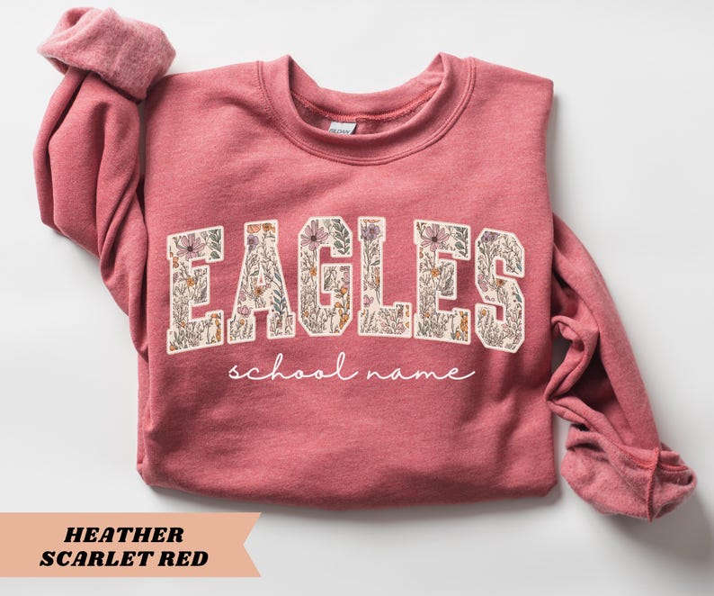 May include: Heather scarlet red sweatshirt with the word "EAGLES" in a floral design. Below the word "EAGLES" is the phrase "school name" in a cursive font. The sweatshirt has long sleeves and a crew neck.