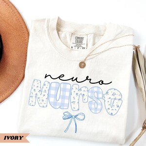 May include: Ivory-colored t-shirt with the words "neuro Nurse" in black script and blue floral and gingham print. A blue bow graphic is below the word "Nurse". A gold necklace with a pendant is on the shirt.