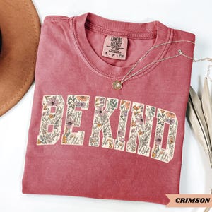May include: Crimson-colored t-shirt with the words "BE KIND" in a floral design. The shirt has a crew neck and short sleeves. A silver necklace with a pendant is draped across the shirt. The shirt is made by Comfort Colors.