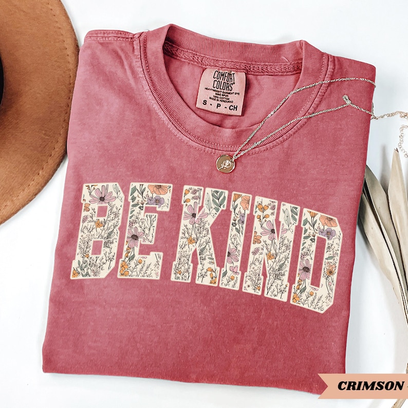 May include: Crimson-colored t-shirt with the words "BE KIND" in a floral design. The shirt has a crew neck and short sleeves. A silver necklace with a pendant is draped across the shirt. The shirt is made by Comfort Colors.