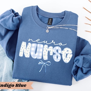 May include: Indigo blue sweatshirt with the words "neuro nurse" in white script and floral and gingham print. A silver necklace with a small pendant is draped across the front. The sweatshirt is folded on top of a pair of blue jeans.