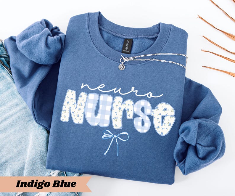 May include: Indigo blue sweatshirt with the words "neuro nurse" in white script and floral and gingham print. A silver necklace with a small pendant is draped across the front. The sweatshirt is folded on top of a pair of blue jeans.