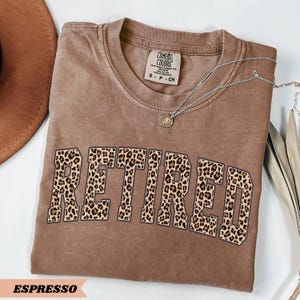May include: Espresso-colored t-shirt with the word "RETIRED" in a leopard print design. The shirt has a crew neck and short sleeves. A silver necklace with a pendant is draped across the shirt. The shirt is folded on a white surface.