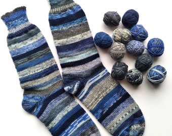 May include: A pair of hand-knitted socks in shades of blue, gray, and white, with a striped pattern. Several small balls of yarn in coordinating colors are arranged nearby. The socks appear to be made of soft wool.