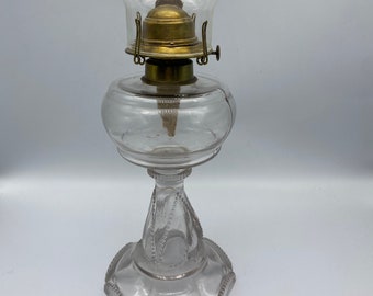 May include: A clear glass oil lamp with a brass burner and a decorative base. The lamp has a round, clear glass globe.