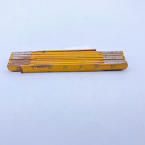 Puede incluir: Una regla plegable de madera amarilla con marcas negras. La regla mide 15,24 cm de largo y tiene marcas para cada pulgada. La regla está hecha en los Estados Unidos.