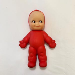 May include: A vintage Kewpie doll with a bald head, rosy cheeks, and a smiling expression. The doll is wearing a solid red onesie. The doll's arms and legs are outstretched, and it has a small red dot on top of its head.
