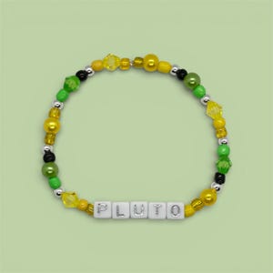 Puede incluir: Una pulsera de cuentas con el nombre "PLUTO" escrito con cuentas de letras blancas. La pulsera presenta cuentas amarillas, verdes, negras y plateadas. Las cuentas son de diferentes formas y tamaños, creando un accesorio colorido.