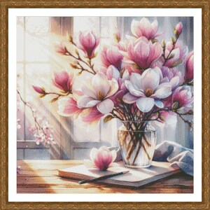 Peut inclure: Un bouquet de fleurs de magnolia roses et blanches dans un vase en verre transparent sur une table en bois avec un carnet et un crayon. Les fleurs sont &eacute;clair&eacute;es par la lumi&egrave;re du soleil qui traverse une fen&ecirc;tre.