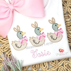 May include: White t-shirt with embroidered ducklings wearing bunny ears and pink bows. The ducks have green heads and yellow beaks. The name "Josie" is embroidered in pink script below the ducks. A large pink bow is in the upper left corner.