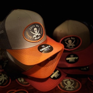 May include: A stack of gray, orange, and red baseball caps with a black mesh back. The caps have a black and white embroidered patch with a skull and crossbones and the text "Calico Jack Apparel". The caps are on a wooden surface with several more patches.