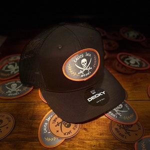 Puede incluir: Una gorra de camionero de malla negra con un parche bordado en negro y naranja que dice "Calico Jack Apparel" y presenta una calavera y huesos cruzados. La gorra está rodeada de varios parches más pequeños con el mismo diseño.