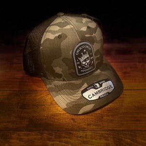 May include: A camouflage trucker hat with a black mesh back. The hat has a black patch with a skull and crown design and the text "Calico Jack Apparel Est. 2021" on it. The hat also has a silver patch with the text "Cambridge Since 2000 PB222".