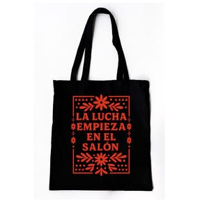 May include: Black tote bag with red floral design and text that reads "LA LUCHA EMPIEZA EN EL SALÓN". The bag has two black handles and is made of a sturdy material. The design is centered on the front of the bag.