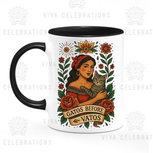 May include: A white ceramic mug with a black handle and rim. The mug features a colorful illustration of a woman holding a cat, surrounded by flowers. The text "GATOS BEFORE VATOS" is displayed on a banner.