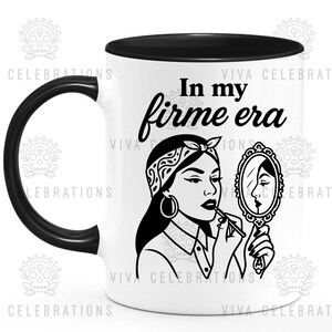 May include: White ceramic mug with a black handle and rim. The mug features a black and white illustration of a woman applying lipstick while looking in a mirror. Text on the mug reads "In my firme era."