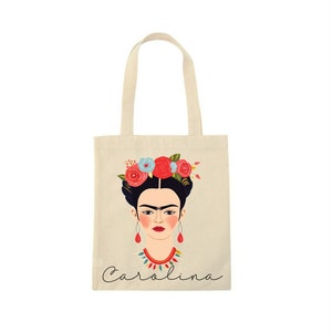May include: A beige tote bag featuring a colorful illustration of a woman with a floral crown, red earrings, and a necklace. The name "Carolina" is written in cursive at the bottom of the bag.