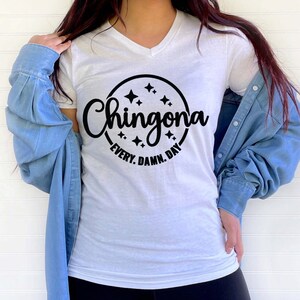May include: White V-neck t-shirt with a black circular design. The design features the word "Chingona" in a script font, surrounded by stars, and the text "Every. Damn. Day." in a smaller font.
