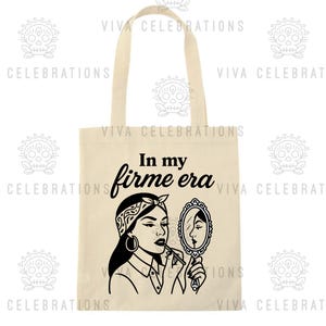 May include: Cream-colored tote bag with a black graphic of a woman applying lipstick while looking in a mirror. The text "In my firme era" is above the graphic. The bag has two handles and is made of a canvas-like material.