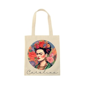 May include: A cream-colored tote bag featuring a portrait of a woman with a floral headdress. The portrait is set within a circular design with colorful flowers. The name "Carolina" is written in cursive below the portrait.