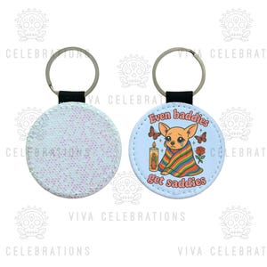 May include: Round keychain with a glittery back and a light blue front. The front features a sad chihuahua wrapped in a rainbow blanket, with butterflies, a candle, and the text "Even baddies get saddies."