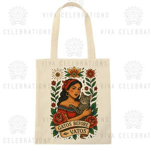 May include: Cream-colored tote bag featuring a woman holding a gray tabby cat, surrounded by floral and leaf designs. The text "GATOS BEFORE VATOS" is displayed on a banner. The bag has a simple handle.