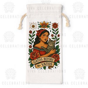 May include: A white canvas wine bag with a drawstring closure. The bag features a colorful illustration of a woman holding a gray tabby cat, surrounded by flowers. The text reads "GATOS BEFORE VATOS".