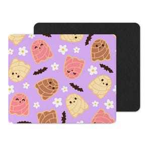 Puede incluir: Una alfombrilla de rat&oacute;n cuadrada con un fondo morado claro que presenta un patr&oacute;n repetido de pasteles de concha sonrientes en colores rosa, marr&oacute;n y crema, junto con flores blancas y murci&eacute;lagos negros. Una almohadilla rectangular negra est&aacute; detr&aacute;s.