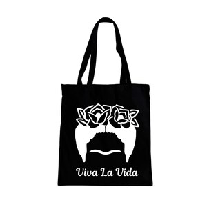 May include: Black tote bag featuring a white graphic of a woman's face with a floral crown and mustache. The words "Viva La Vida" are printed in white below the graphic. The bag has black handles.