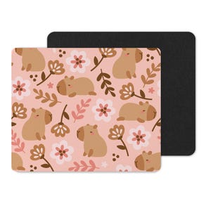 Op de afbeelding: Een rechthoekige muismat met een herhalend patroon van cartoon capibara's, bloemen en bladeren in bruin, roze en wit. De mat heeft een zwarte achterkant.