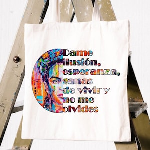 Puede incluir: Un bolso tote de lona blanca con un retrato colorido de una mujer y texto en español: "Dame ilusión, esperanza, ganas de vivir y no me olvides." La obra de arte es una vibrante mezcla de colores.