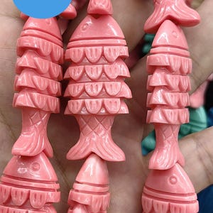 Op de afbeelding: Een streng roze koraal kralen in de vorm van vissen. Elke vis kraal heeft een gedetailleerd gesneden ontwerp van schubben en vinnen. De kralen zijn aan elkaar geregen op een doorzichtige draad.