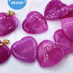 May include: A collection of heart-shaped pendants in varying shades of magenta. Some pendants have gold-colored bails. The pendants are smooth and glossy, with a marbled appearance. The word "BEADS" is in a blue circle in the upper left corner.