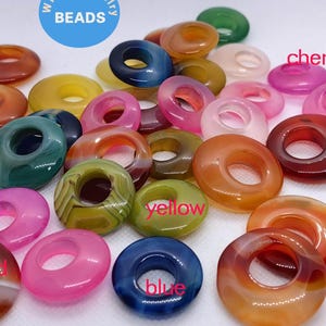 May include: Assortment of colorful agate donut beads with holes in the center. The beads are in shades of green, red, yellow, blue, and brown. The beads are labeled with the colors: green, red, yellow, blue, cherry.