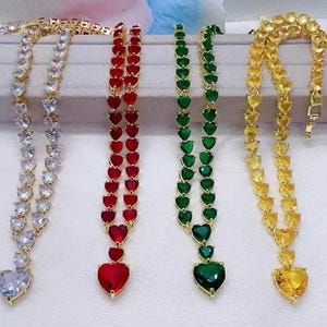 May include: Four gold heart-shaped necklaces with different colored gemstones. The necklaces feature a row of smaller heart-shaped stones and a larger heart-shaped pendant. The colors of the gemstones are white, red, green, and yellow.