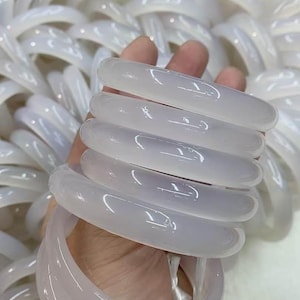 Puede incluir: Un conjunto de brazaletes de jade blanco, también llamados bangles, se sostienen en una mano. Los brazaletes son lisos y pulidos.