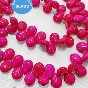 Puede incluir: Un conjunto de cuentas en forma de lágrima en tonos rosa y magenta vibrantes, dispuestas sobre una superficie blanca. Se observa un círculo azul con la palabra "BEADS". Estas cuentas son probablemente para la creación de joyas.