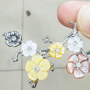 May include: A silver-toned floral brooch featuring a branch design with several flower blossoms. The flowers are in shades of white, yellow, and pink, each with a central crystal. The brooch is held by a person's fingers.