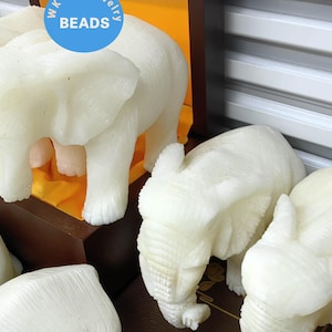 Può includere: Cinque statuette di elefante in alabastro bianco in mostra. Gli elefanti sono disposti in fila su uno scaffale di legno con uno sfondo di tessuto giallo. Tutti gli elefanti sono rivolti nella stessa direzione e hanno la proboscide alzata.