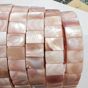 Puede incluir: Primer plano de una pulsera hecha de cuentas rectangulares, iridiscentes, de nácar rosa y blanco. Las cuentas están dispuestas en una fila, creando un accesorio flexible y llamativo. La pulsera es una hermosa pieza de joyería.