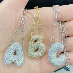 Puede incluir: Tres collares iniciales, cada uno con una letra diferente: A, B y C. Las letras están incrustadas con pequeñas piedras brillantes. Las cadenas son plateadas y doradas, con un estilo de clip.