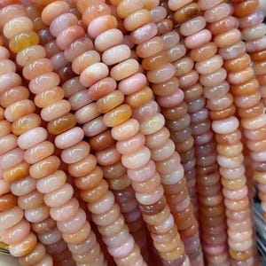 Pode incluir: Várias fileiras de contas de pedras preciosas em tons de pêssego, rosa e creme. As contas são redondas e polidas. A imagem mostra várias fileiras, sugerindo que estão prontas para fazer joias.