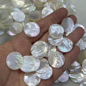 May include: A collection of round, white mother of pearl shell cabochons with iridescent rainbow colors. The cabochons are approximately 1 inch in diameter.