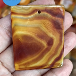 Op de afbeelding: Een vierkante agaat hanger met een gat geboord aan de bovenkant. De hanger heeft een bruin, geel en oranje kleurenschema met een draaiende patroon.