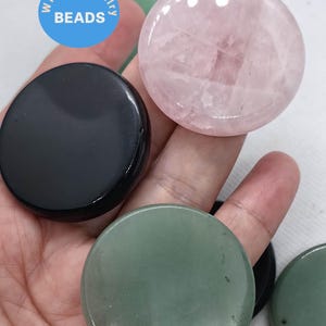 May include: A hand holds several polished, round gemstone beads in various colors. The beads include black onyx, rose quartz, and green aventurine. The image also includes a blue circle with the text "WK DIY Jewelry BEADS."